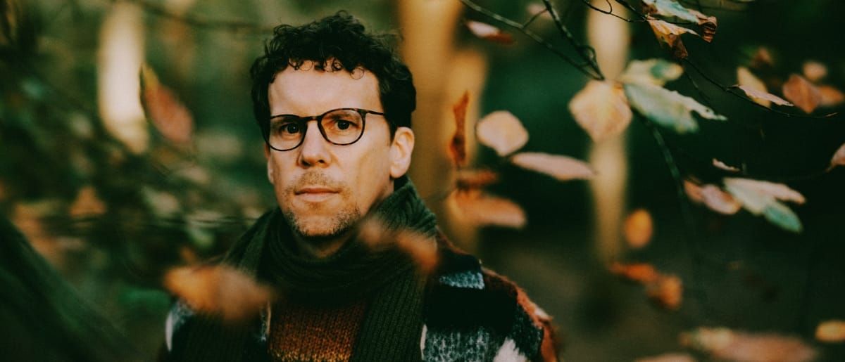 Scrimshire photographed among autumn woodland foliage, wearing glasses and a patterned knit sweater, looking directly into the camera.