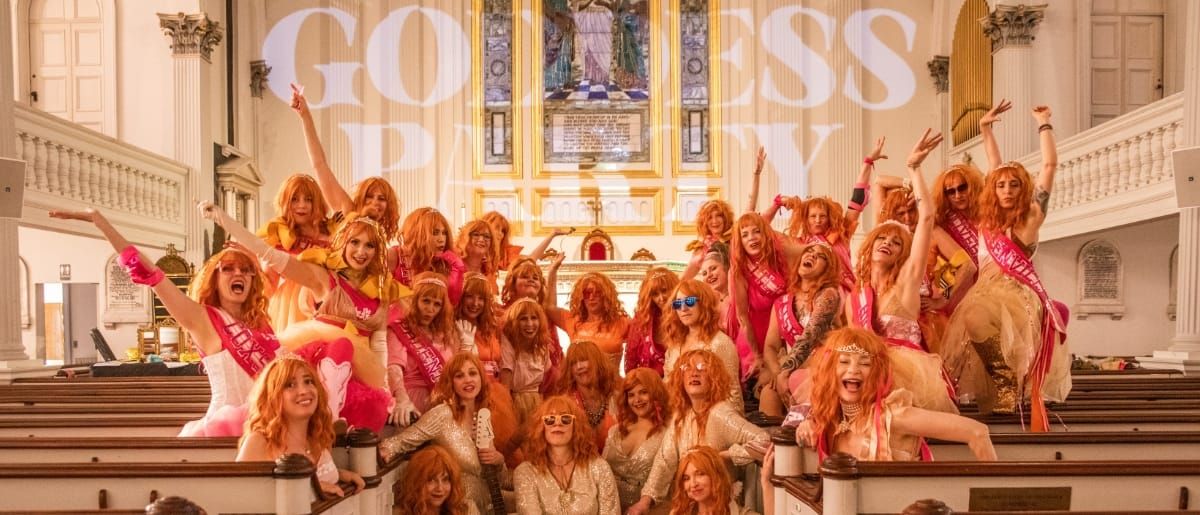 Dozens of The Goddess Party performers in orange wigs and pink and gold sequins pose across the pews of an ornate church, arms raised, 'GODDESS PARTY' projected on the wall behind them.