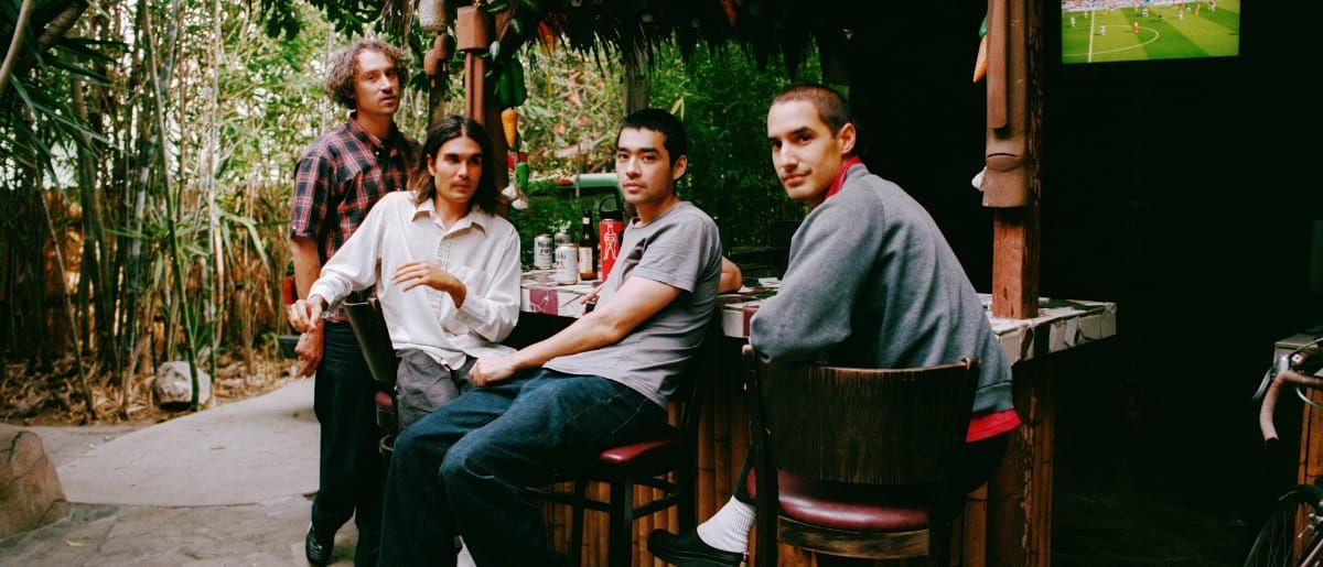 Bondo sit at an outdoor bamboo bar ringed with tropical plants, all four turning to face the camera, a soccer match playing on a screen above.