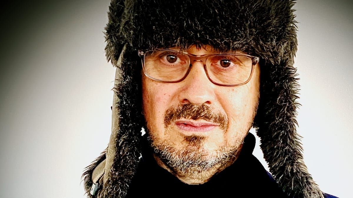 David Norland in a thick fur trapper hat with ear flaps and clear-framed glasses, looking directly at the camera against a pale grey wall.