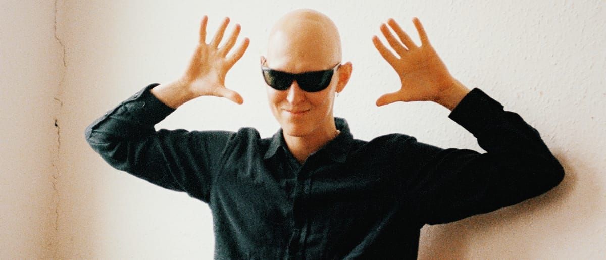 GB stands against a white wall in a black shirt and dark sunglasses, both hands raised palms-out beside his head.