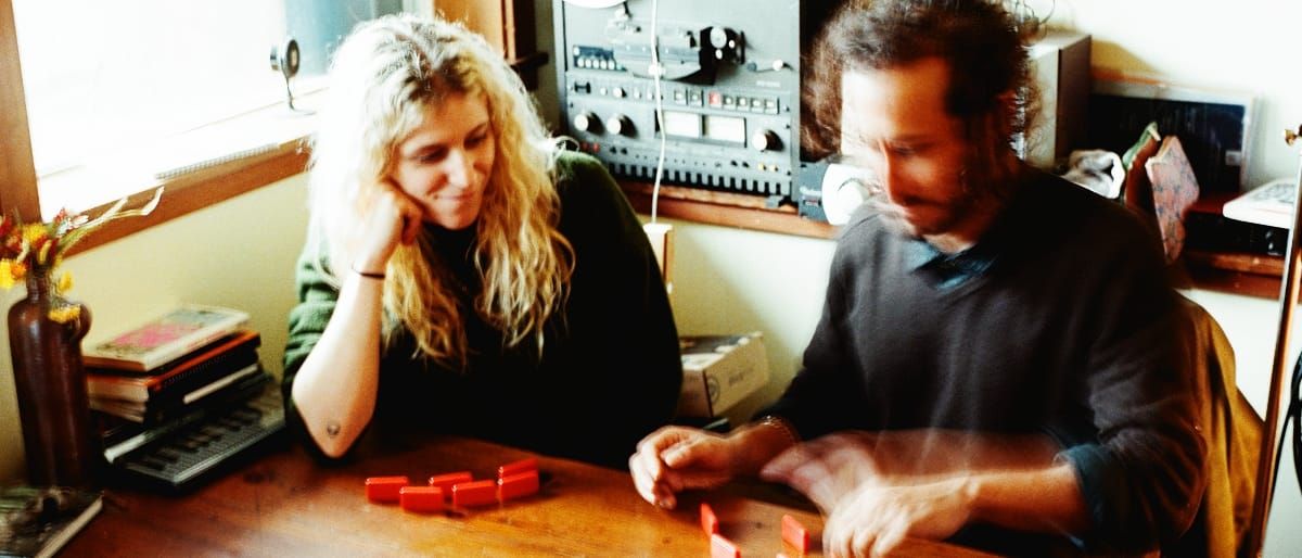 Julia Shapiro rests her chin on one hand, watching Samur Khouja arrange small red tiles on a wooden table, his hands blurred with movement, a reel-to-reel tape machine behind them.