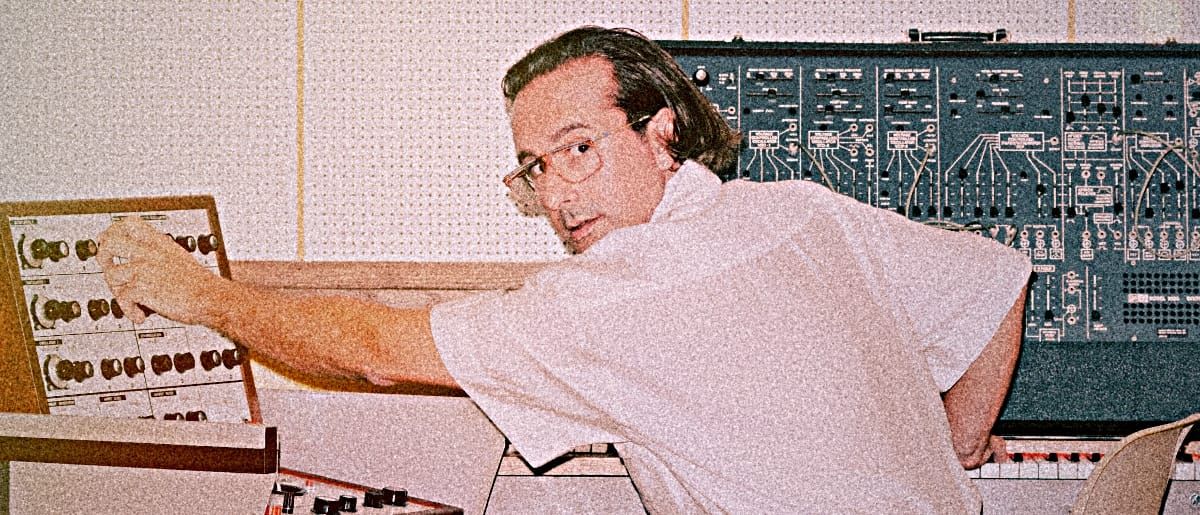 Jolly Mare, in glasses, reaches to turn a knob on a mixing panel, a large modular synthesizer mounted on the wall behind him.