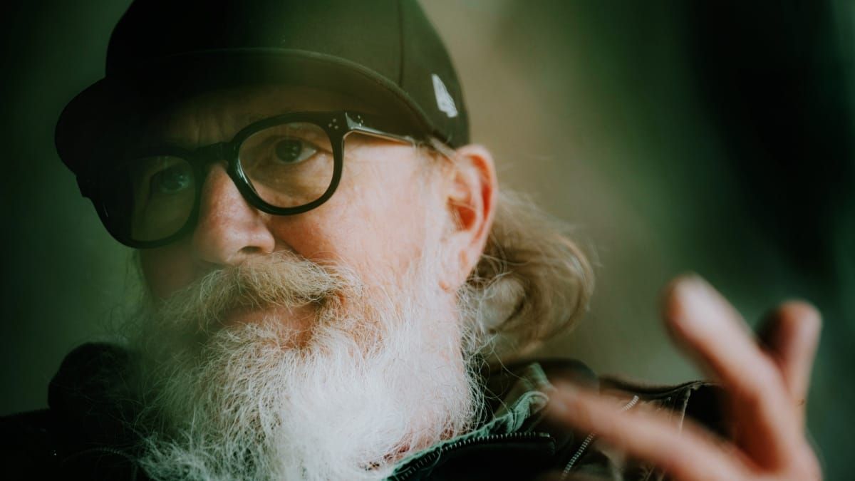 Close-up of Kurt Wagner in a black cap and heavy black-framed glasses, his full white beard filling the frame, one hand raised and blurred in the foreground against soft green shadow.