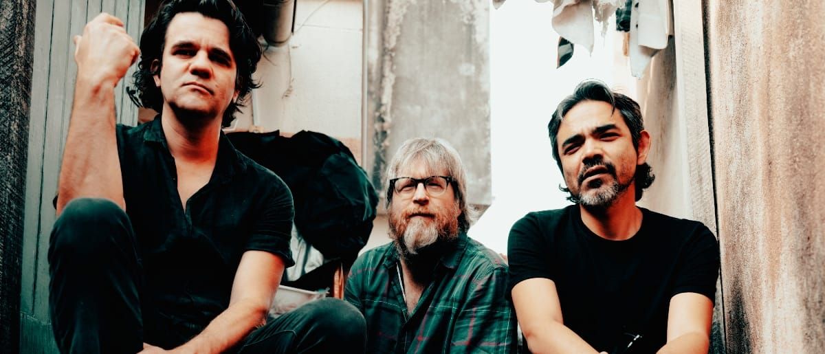 Riviera Gaz seated in a narrow alley with peeling walls — Gustavo Riviera at left with a raised fist, Steve Shelley centre in a plaid shirt, Paulo Kishimoto at right in a black T-shirt.