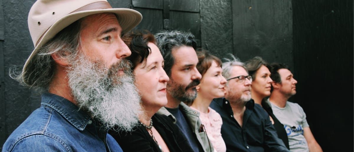 The Odyssey Cult in a row against a black wooden wall, all in profile facing right, Ethan Miller nearest the camera in a pale hat and denim jacket, long grey beard.