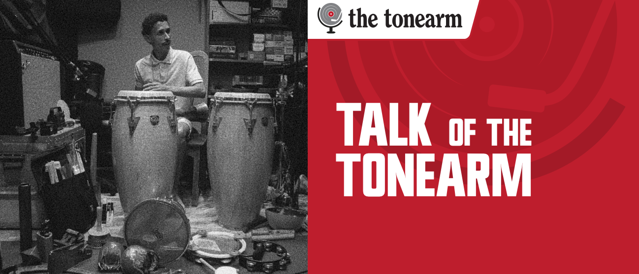 Irán Farías Saínz sits behind two congas in a cluttered studio ringed with tambourines, shakers, and cowbells. A red panel beside him reads TALK OF THE TONEARM.
