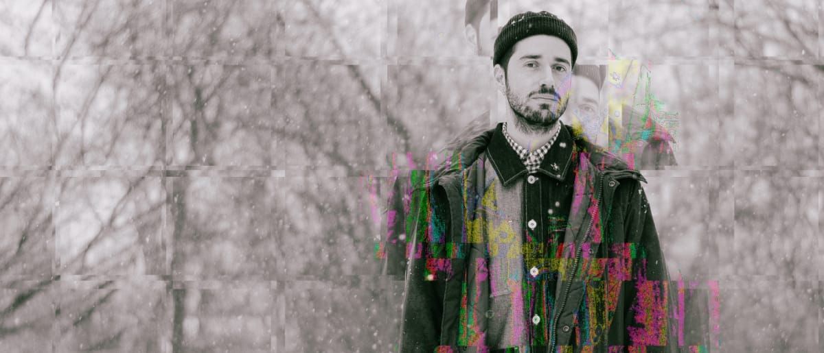 arc rae stands in falling snow before bare winter trees, wearing a knit cap and dark jacket, the image fractured by grid glitches and bursts of neon color across his chest.
