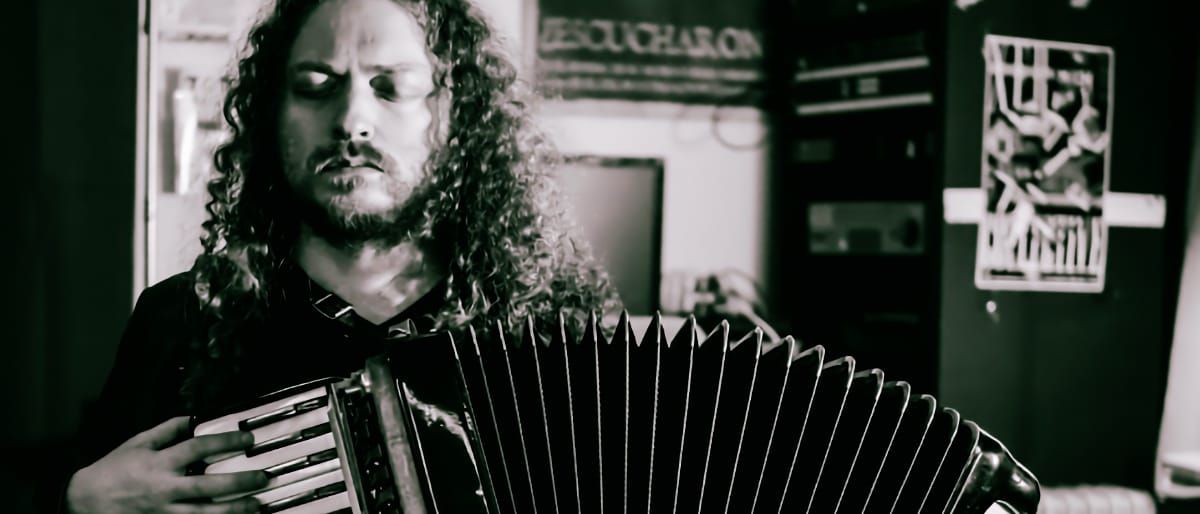 Ben Richter plays accordion in black and white, bellows pulled wide, eyes closed, posters and shelving on the wall behind him.