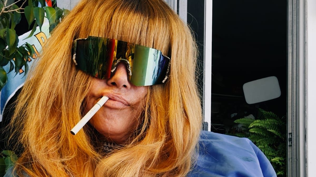 Close-up of Jennifer Herrema in mirrored shield sunglasses, a cigarette in her mouth and long strawberry-blond bangs framing her face.