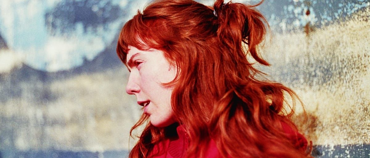 Elanor Moss in profile, red hair pulled into a high ponytail with a fringe, wearing a red top, lips parted. Behind her a blurred backdrop of pale blue and weathered cream.