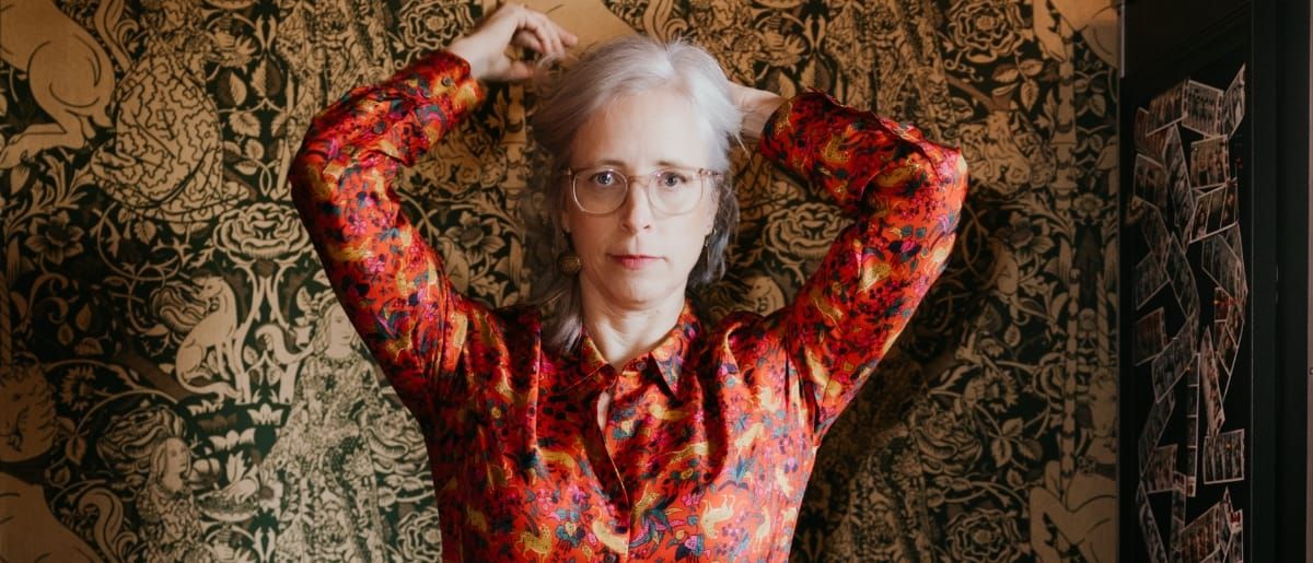 Laura Veirs stands with both arms raised and hands behind her head, looking straight ahead, in a red floral shirt against ornate olive-and-gold figured wallpaper.