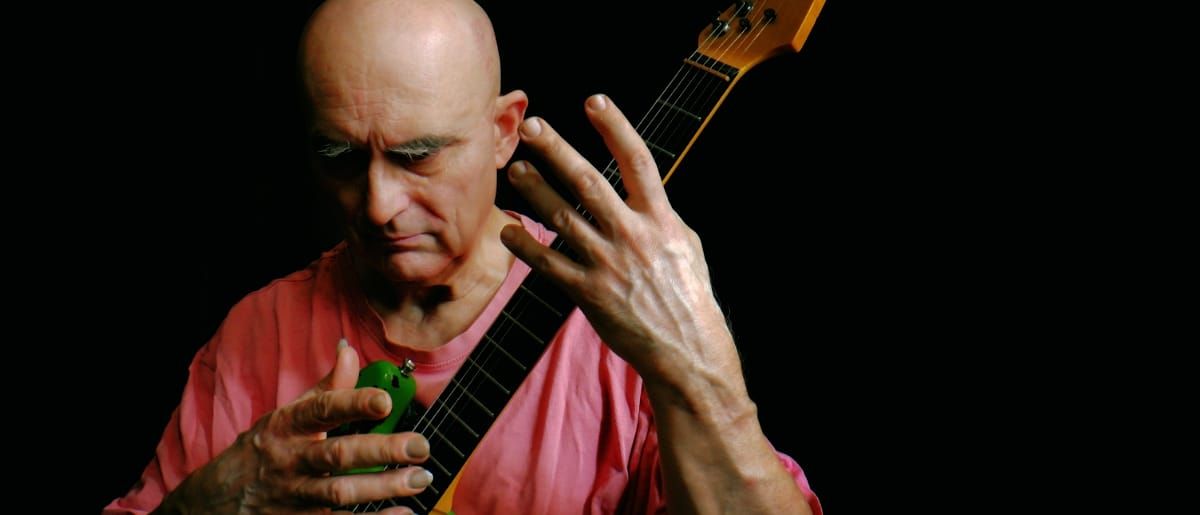 Olaf Rupp bends over an electric guitar held upright against a black background, fingers spread wide across the neck, in a faded pink T-shirt.