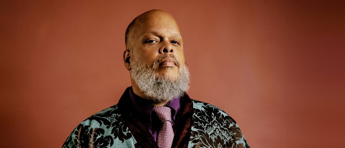 Ed Motta before a terracotta backdrop in a teal and black brocade jacket, purple shirt and pink patterned tie, chin raised, looking into the camera