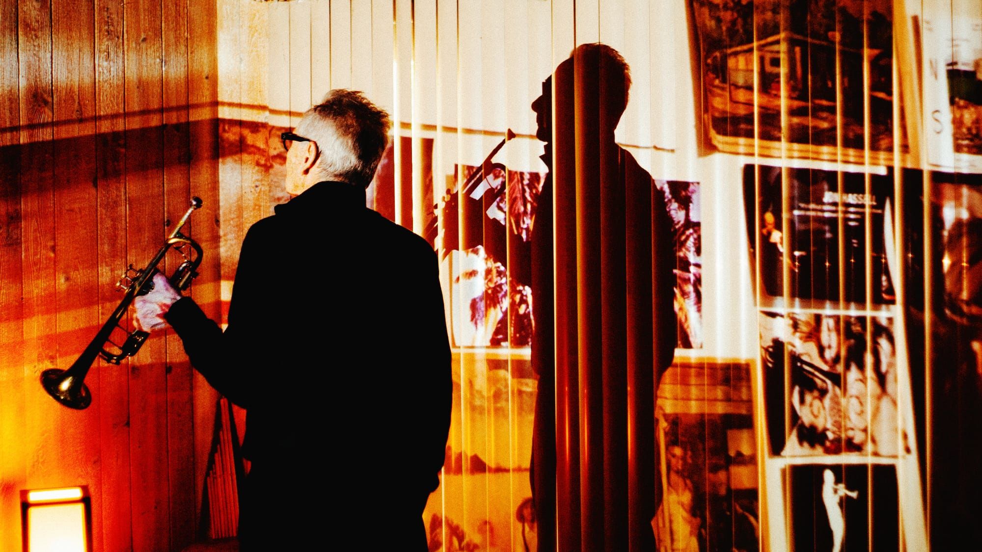 Jon Hassell, seen from behind with trumpet in hand, faces a wall of photographs and posters, his shadow cast large across it through vertical blinds in warm amber light.