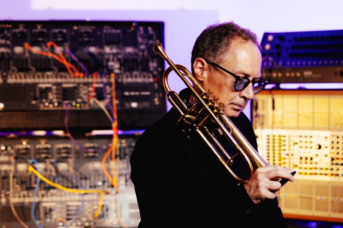 Mark Isham looks down, his trumpet held upright against his shoulder, in front of vintage synthesizers strung with orange and yellow patch cables.