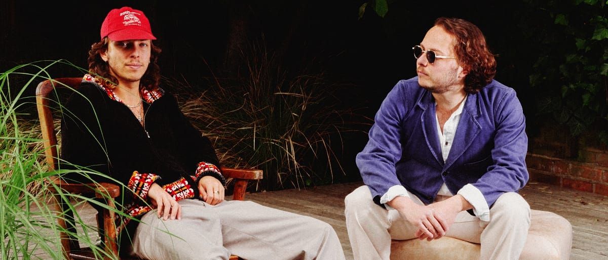 Henry and Reuben Vaun Smith of Sons of Sevilla sit side by side on a dark patio, one in a red cap and patterned black jacket, the other in a blue-violet jacket and sunglasses, looking left.