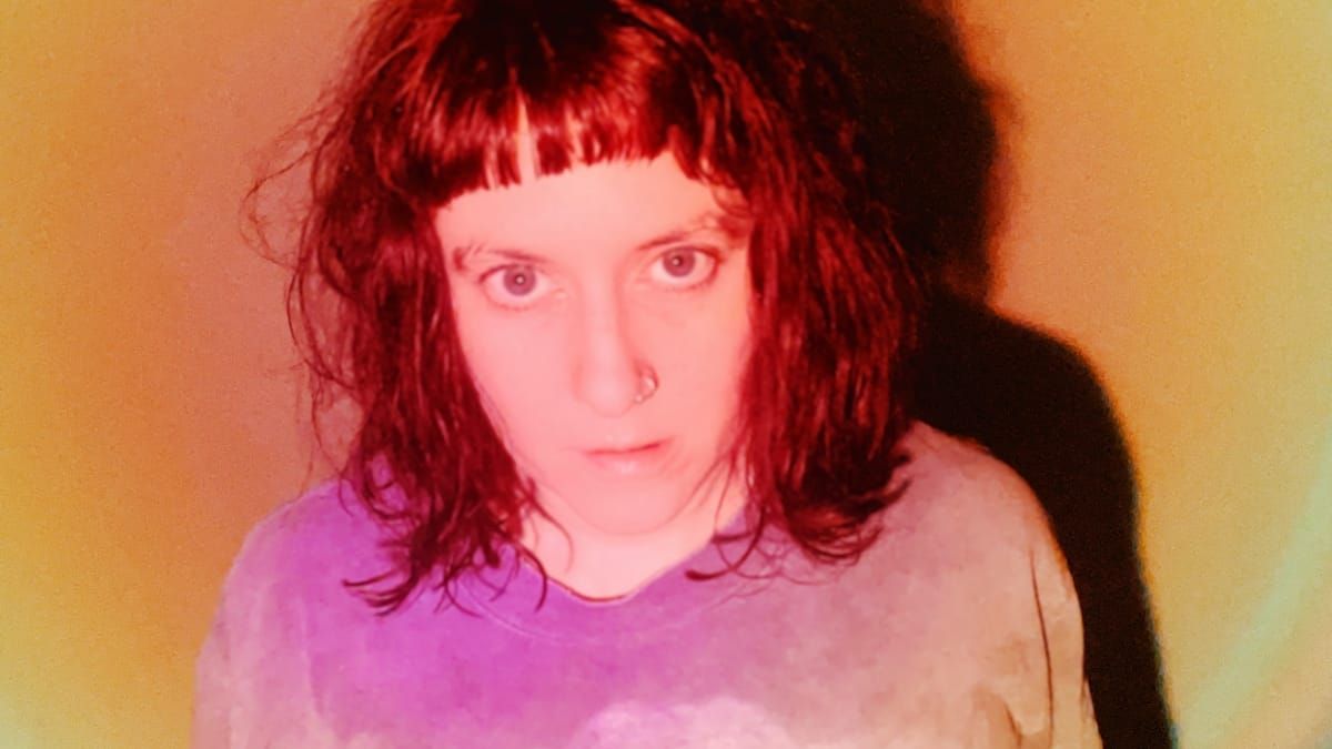 Tiiva in close-up, dark fringe and a nose ring, looking straight at the camera against a warm orange glow, a hard shadow cast behind them.