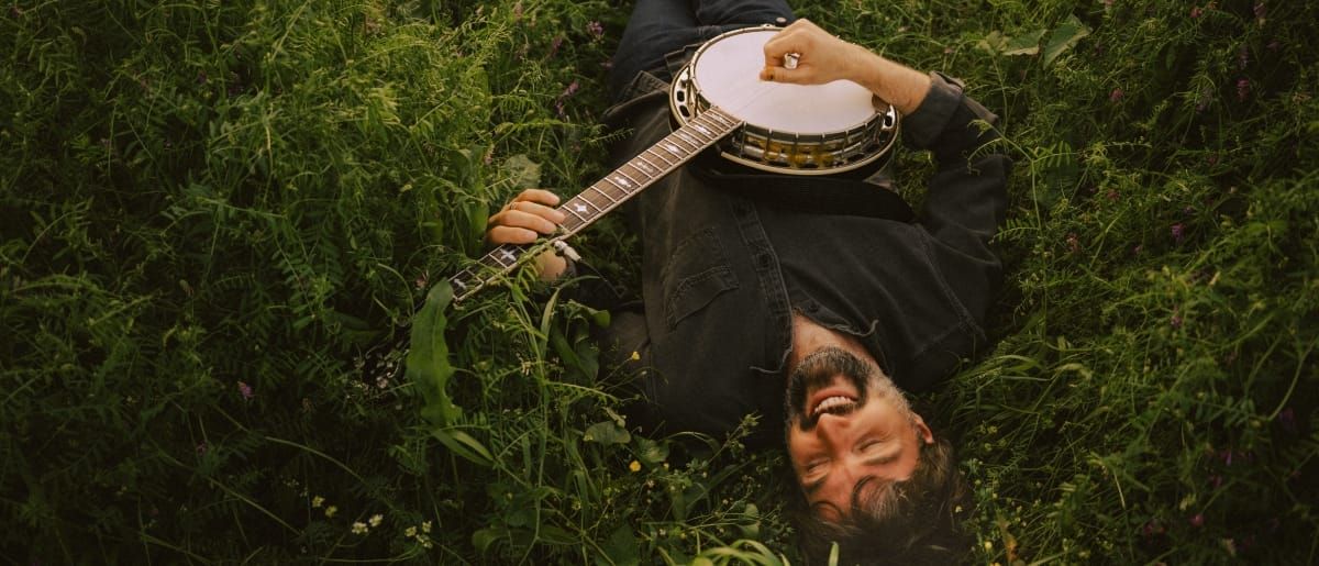 A smiling Wes Corbett playing banjo while lying on the ground. Photo by Hannah Gray Hall.
