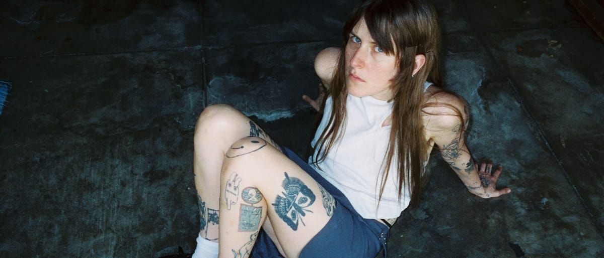 Lala Lala sits on dark textured surface wearing overalls and white turtleneck, displaying visible tattoos on arms and legs. Photo by Ariel Fish.