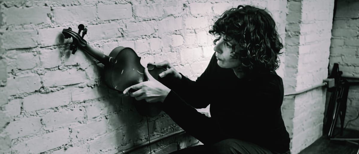 Marija Kovačević tunes a violin against white brick wall in black and white photograph, curly dark hair visible. Photo by Peter Gannushkin.