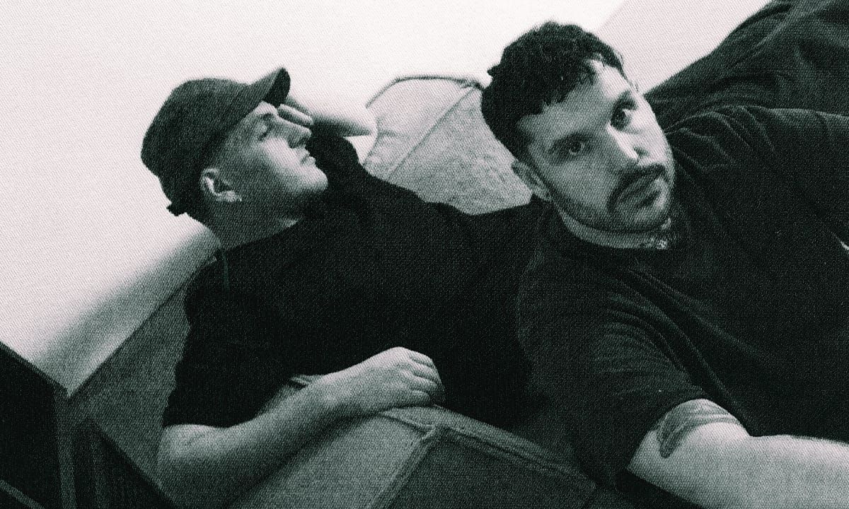 Max Scholl and Dhanya Langer of musical duo Modha photographed from below in black and white. Scholl wears facial hair, Langer wears a cap.