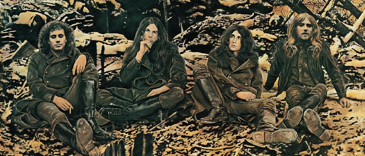 Four members of Armageddon seated on rocky debris in earth-toned vintage photo, wearing leather jackets and boots in casual 1970s style.