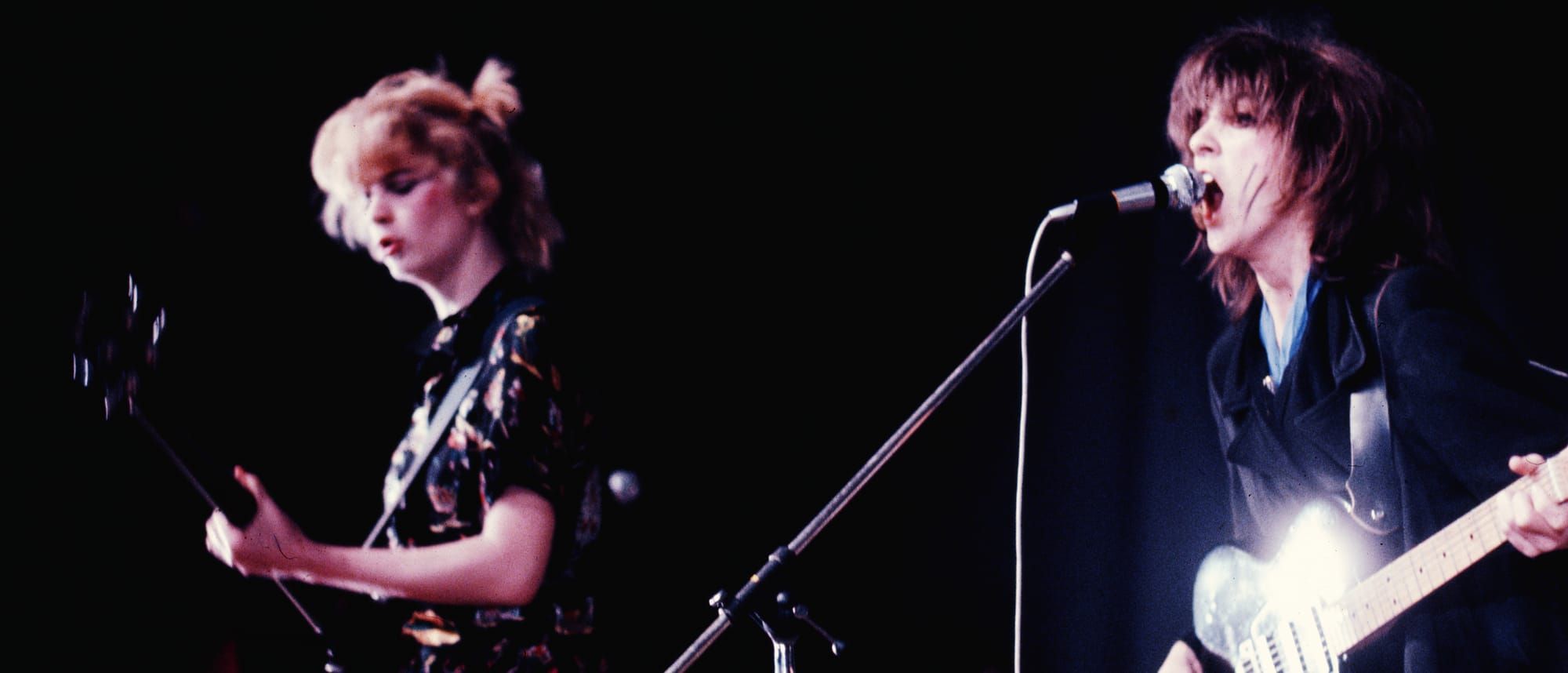 Gina and Ana of The Raincoats perform in Warsaw, 1978, vintage concert photo with guitar and microphone, purple-pink tones. Photo by Maria Helena da Silva and courtesy Ana da Silva.
