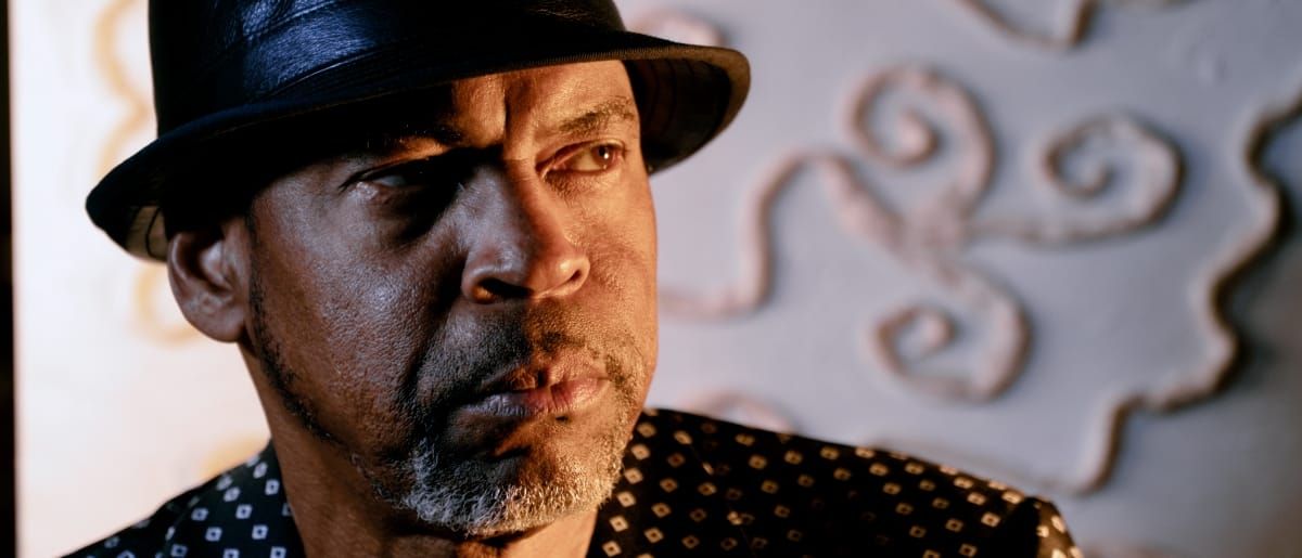 Bayeté (Todd Cochran) in hat and patterned shirt with warm dramatic lighting, decorative scrollwork visible in background.