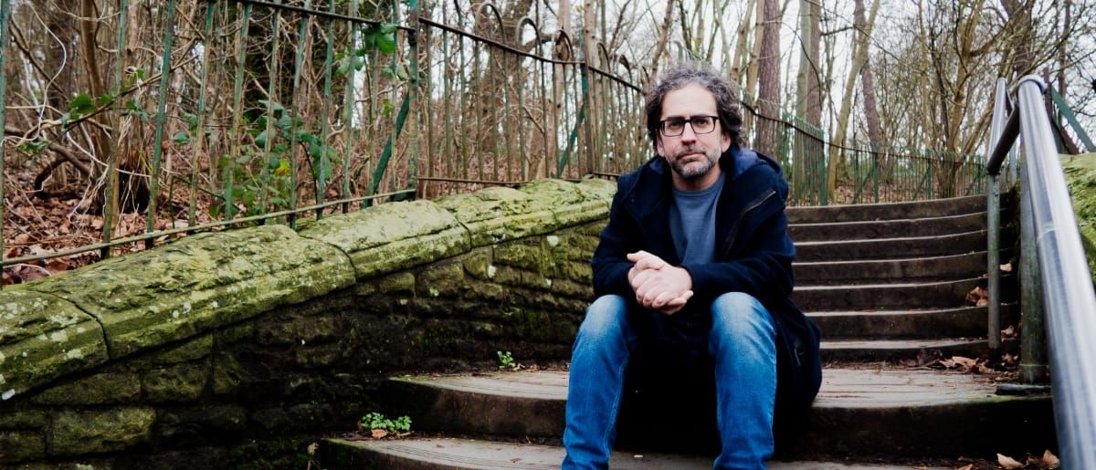 Billy Fuller sits on moss-covered stone steps with hands clasped, wearing a dark coat and jeans, surrounded by bare winter trees and iron railings. Photo by Charlie Romijn-Barr.