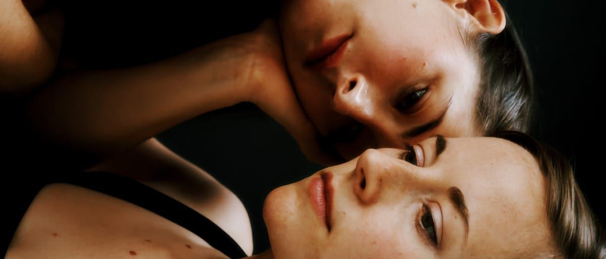 Two members of Sibyl lie close together with warm dramatic lighting, faces near, black clothing visible. Photo by Gregory Wikstrom.