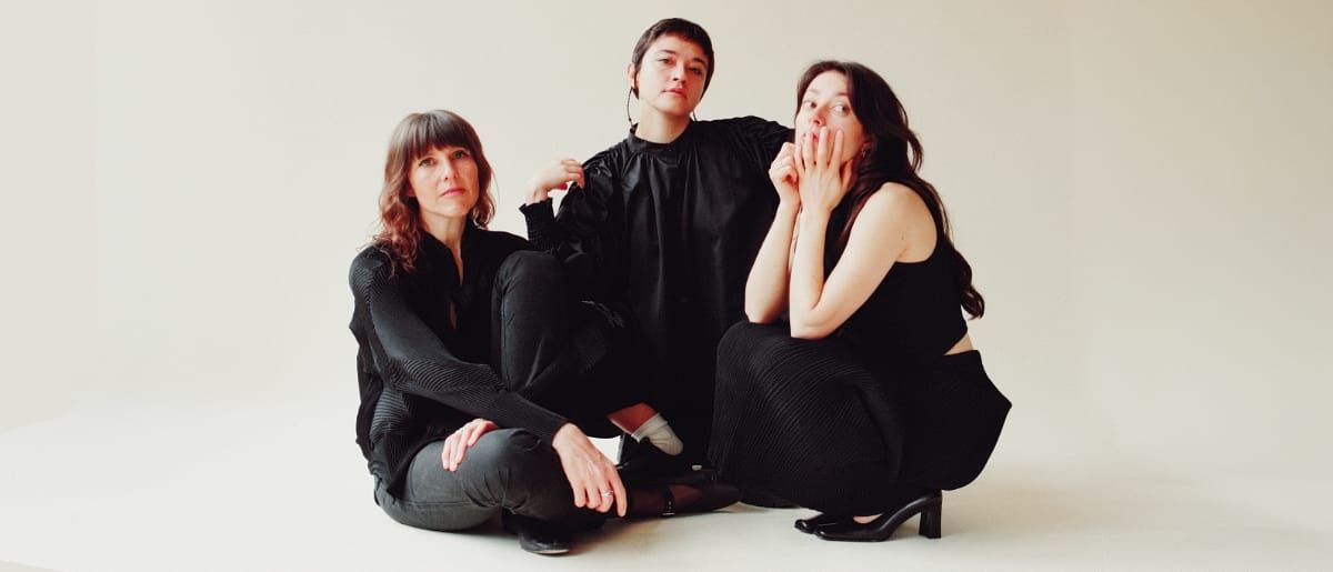Whitney Johnson, Macie Stewart, and Lia Kohl sit together on a white floor in all-black clothing against a cream background, looking directly at the camera. Photo by Leah Wendzinski.