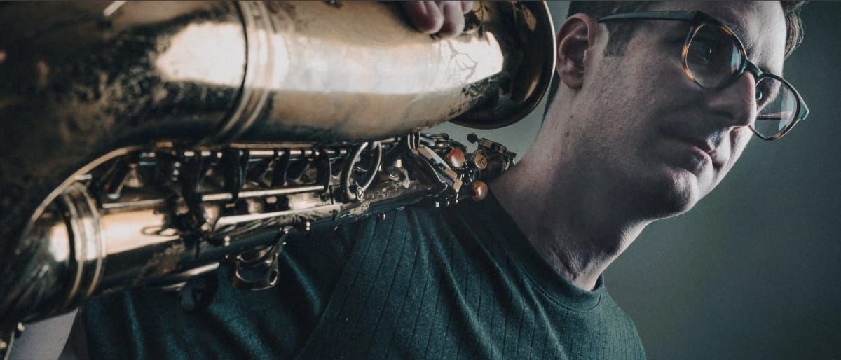 Ethan Helm holds a saxophone resting on his shoulder, looking slightly off-camera. Close crop, muted teal background. Tortoiseshell glasses.