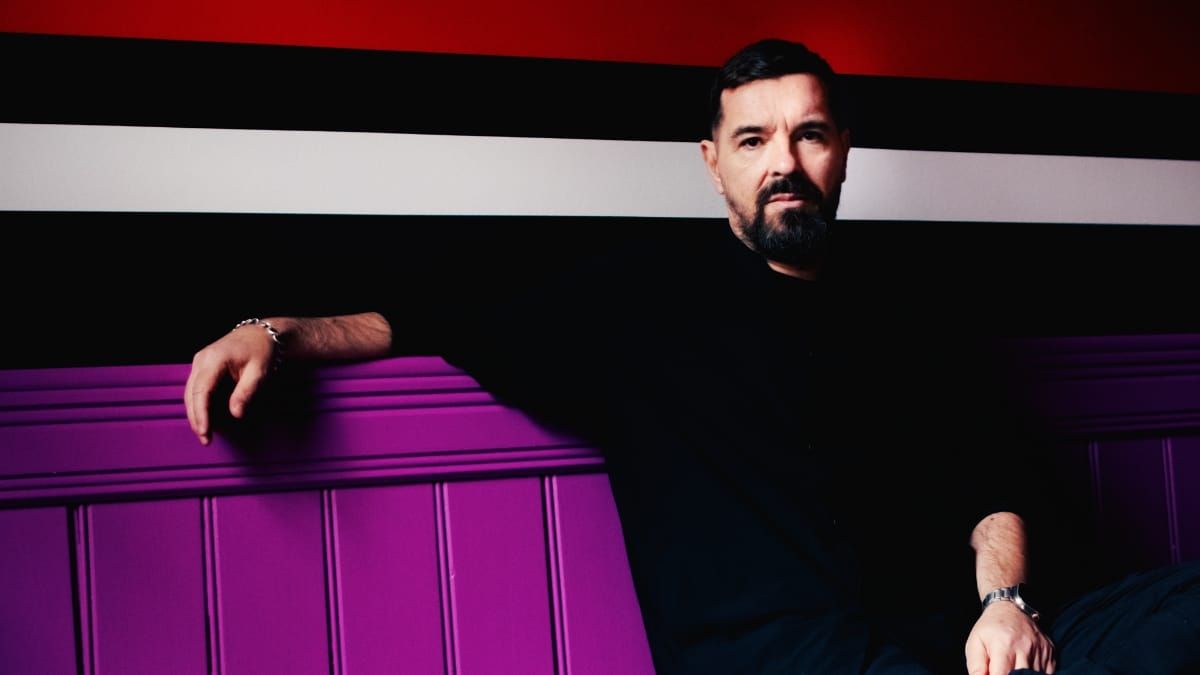 Speedy J leans against a bold purple panel, one arm draped over the top, wearing a black shirt, set against a striped red, black, and white background. Photo by Tommy Spring.