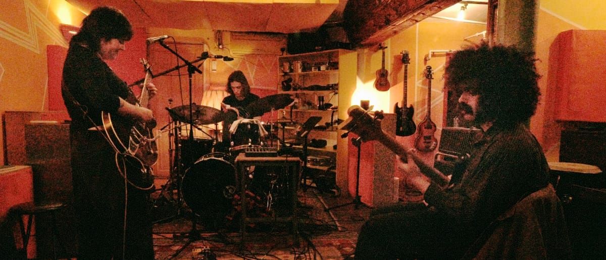 Myra Lee rehearse in a warmly lit studio, guitarist and bassist flanking a drummer at center, guitars hanging on the wall behind them.