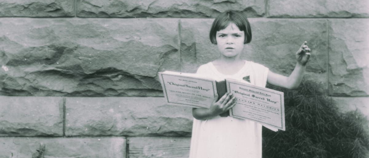 Lorraine Miles holds open copies of the "Original Sacred Harp" hymnal before a stone wall, one arm raised, in a vintage black-and-white photograph.