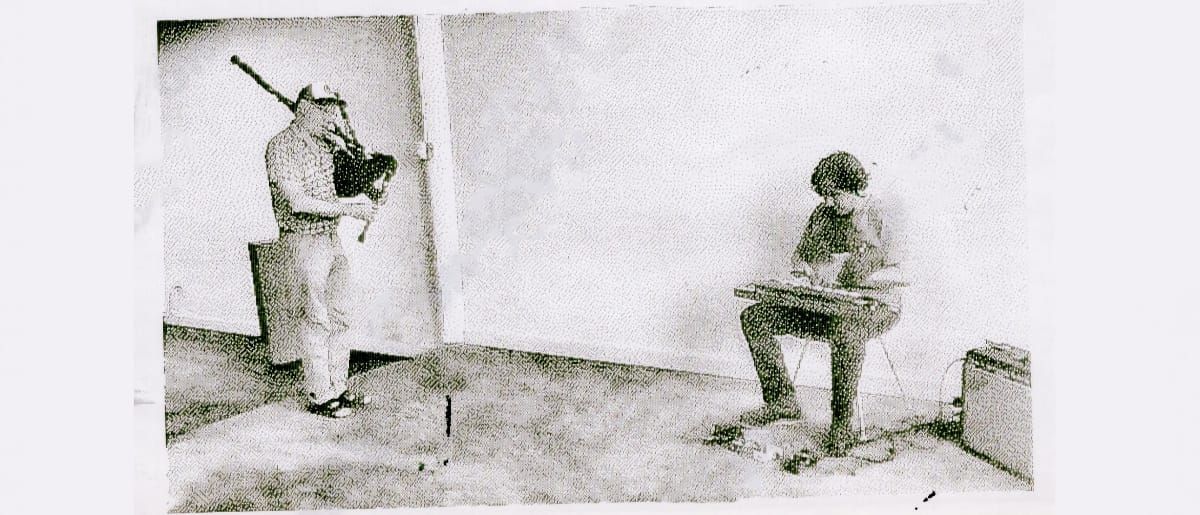 David Watson stands playing Highland bagpipes near a corner, while Bill Nace sits across the room playing a taishōgoto on his lap, in a grainy halftone photograph.