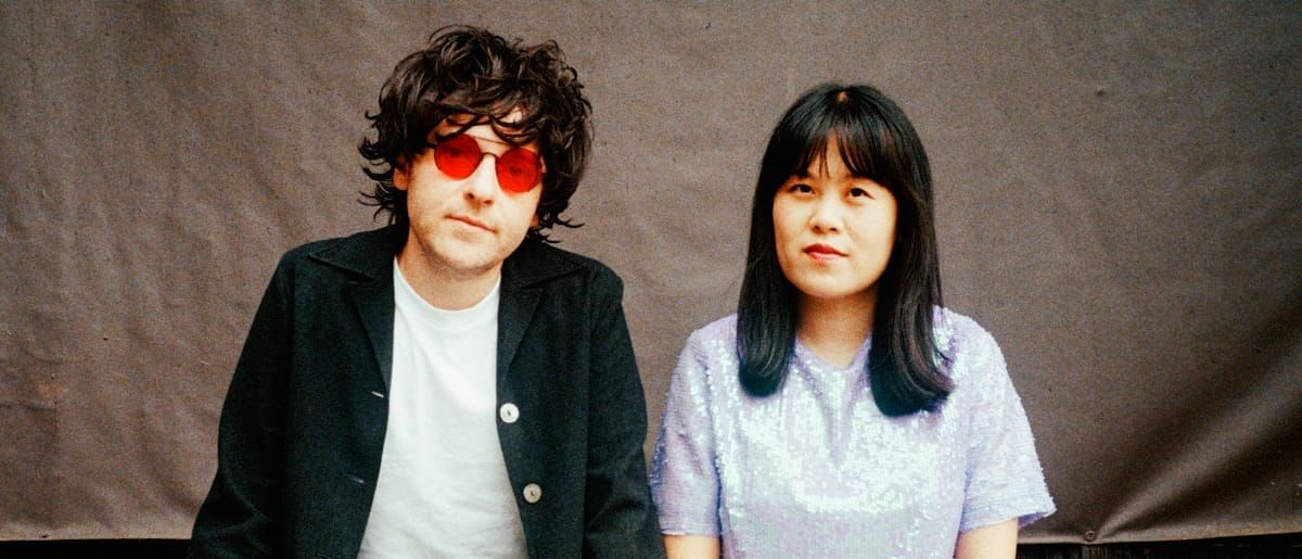 Bryce Barsten and Anne Tong of Chinese American Bear seated against a grey backdrop, Barsten in red tinted round sunglasses and a black jacket, Tong in a lavender sequined top.