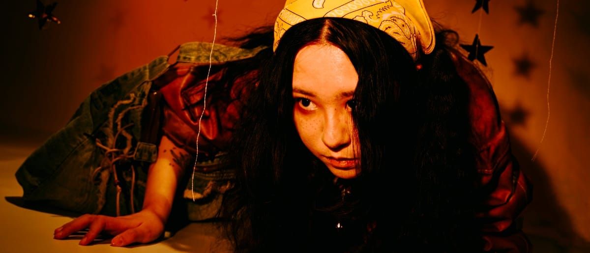 Mixol crawls toward the camera along the floor, hair falling over one eye, in a denim jacket and yellow patterned headscarf, against an orange wall hung with dark stars.