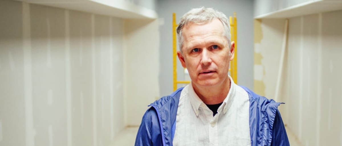 Matthew S of Myth Math stands in an unfinished room, drywall seams exposed and a yellow ladder behind him, wearing a blue windbreaker over a pale button-up shirt, looking at the camera.