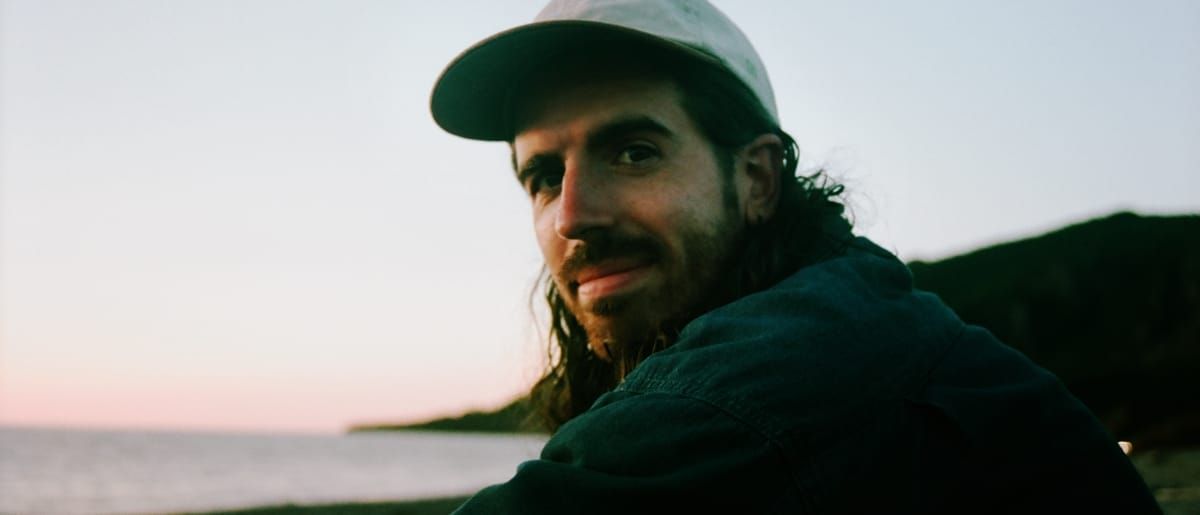 St. Silva glances back over one shoulder in a pale ball cap and denim jacket, a calm sea and dusky pink sky behind him.