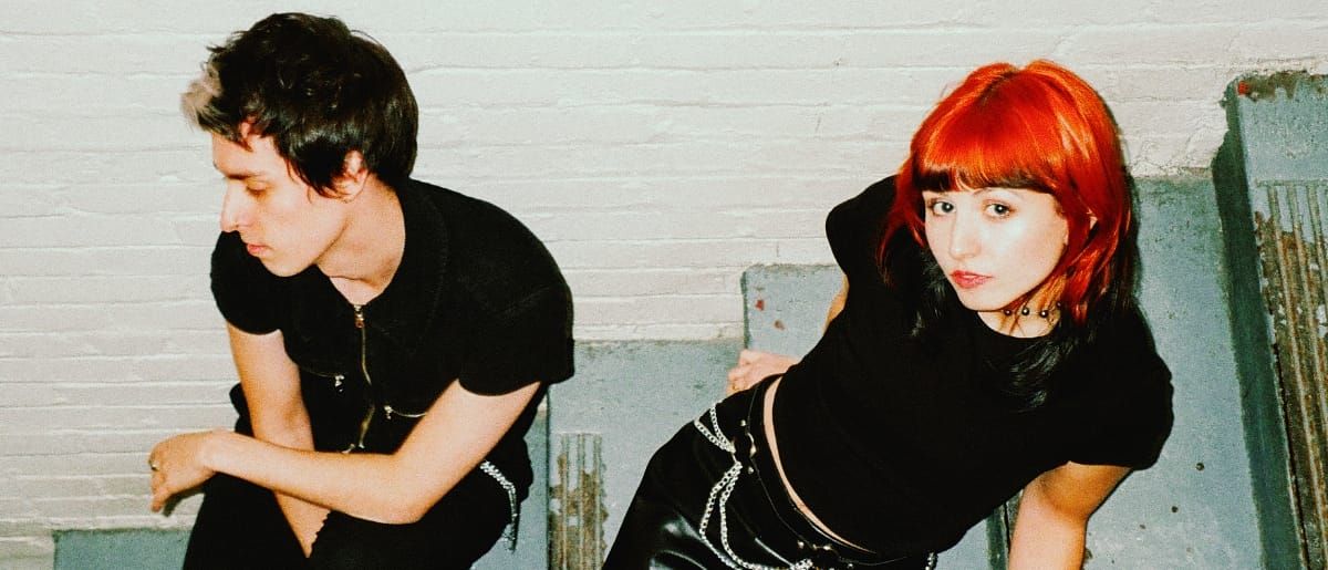 Nikki St. Pierre and Courtney Carmichael of sundayclub lean toward the camera against a brick and metal wall, both in black, Carmichael with vivid red hair.
