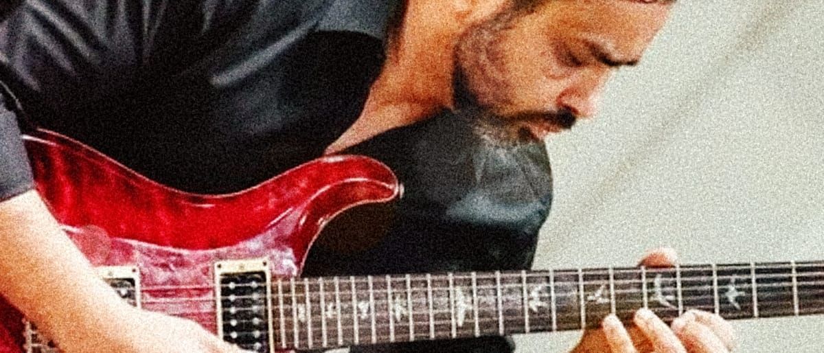 Ayman Fanous bends low over a red electric guitar, eyes on the fretboard as his fingers press the strings.