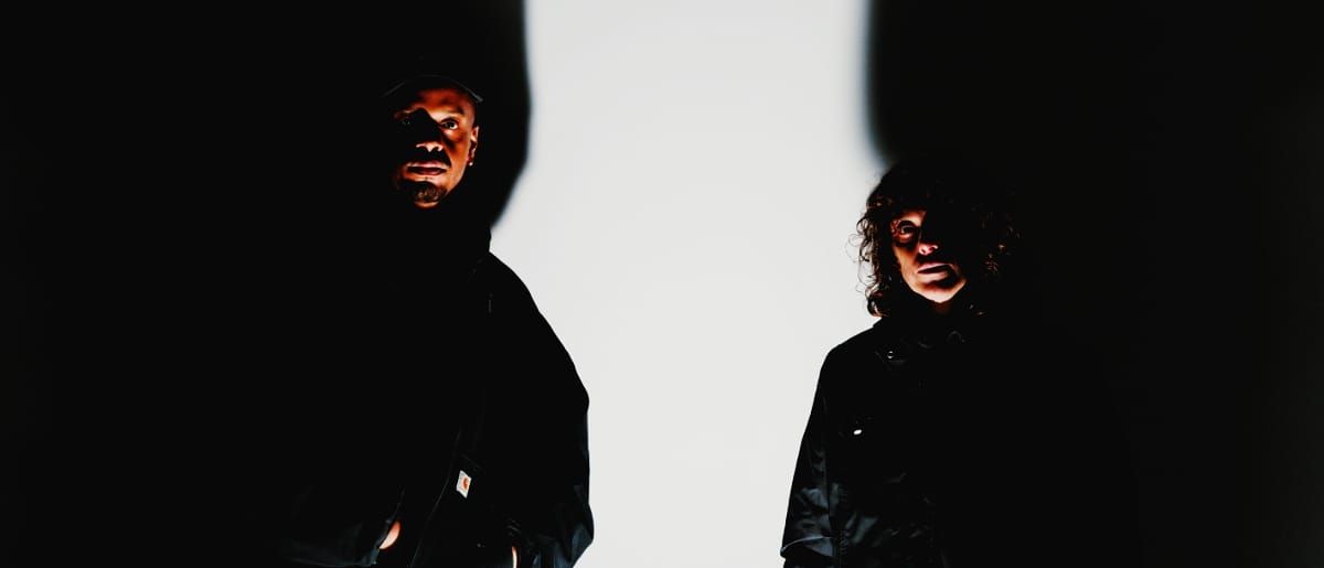 Chris Ryan Williams and Ka Baird look into the camera, faces half-lit in dark clothing, a strip of bright white wall between them.