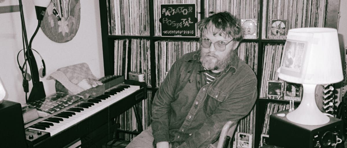 Sam Cook-Parrott in tinted glasses sits slouched between a keyboard and a white table lamp, record shelves and a Radiator Hospital sign behind him. Black and white.