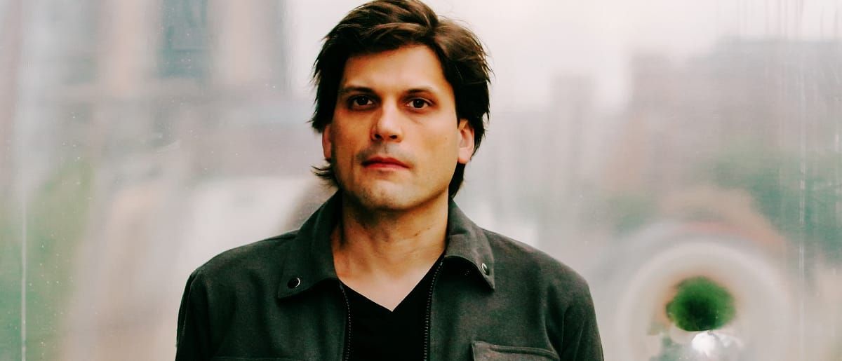Brett Bullion of deep glens faces the camera in a dark jacket, city buildings blurred behind him through a misted window.