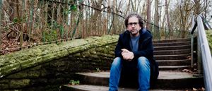 Billy Fuller sits on moss-covered stone steps with hands clasped, wearing a dark coat and jeans, surrounded by bare winter trees and iron railings. Photo by Charlie Romijn-Barr.