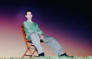 A young man (Gregory Uhlmann) in a green striped shirt and light jeans sits in a wooden folding chair on grass against a twilight gradient sky. Photo by Joyce Kim.