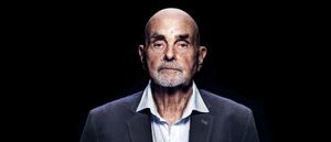 A stark studio portrait of Hans-Joachim Roedelius, bald and white-bearded in a grey blazer and open-collar shirt, staring directly into the camera against a pure black background.
