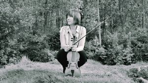 Meredith Bates crouches on grass before a wooded backdrop, holding a violin and bow, gazing to one side.